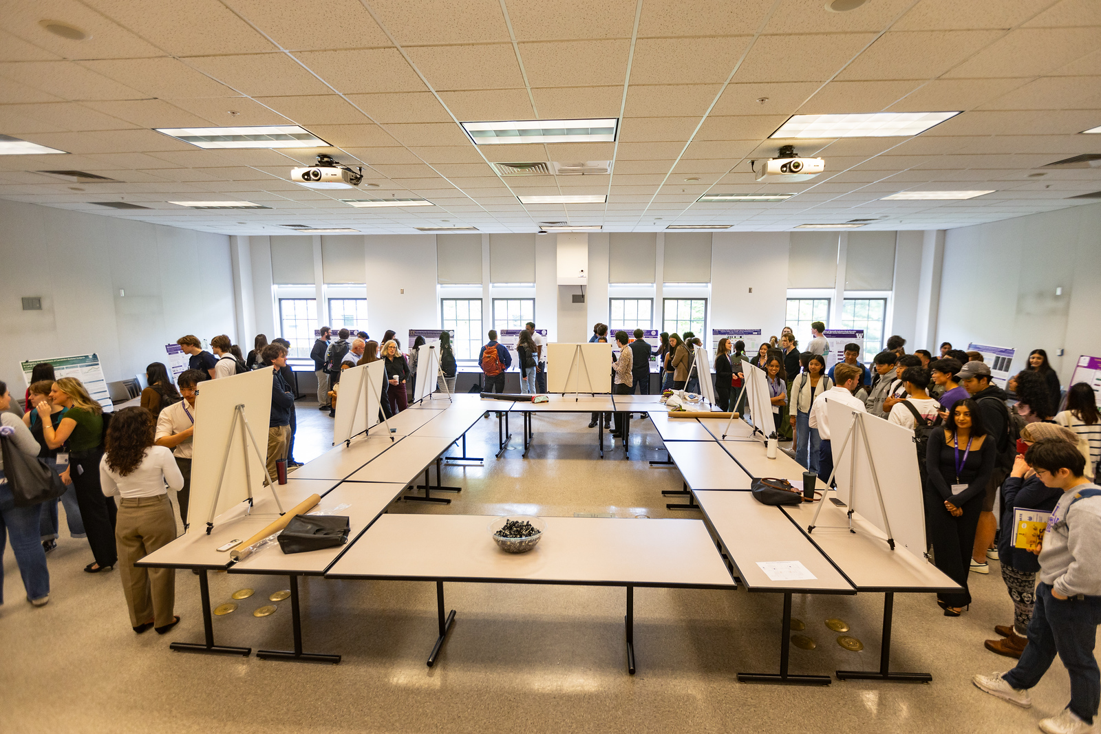 "A photo of a lively event, where people in business attire view posters lined up on rows of tables. Students, the creators of the posters, explain to the business people what the posters represent."