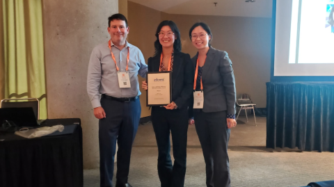 Three conference attendees pose together indoors, with the person in the center holding an INFORMS award certificate and all wearing conference badges.