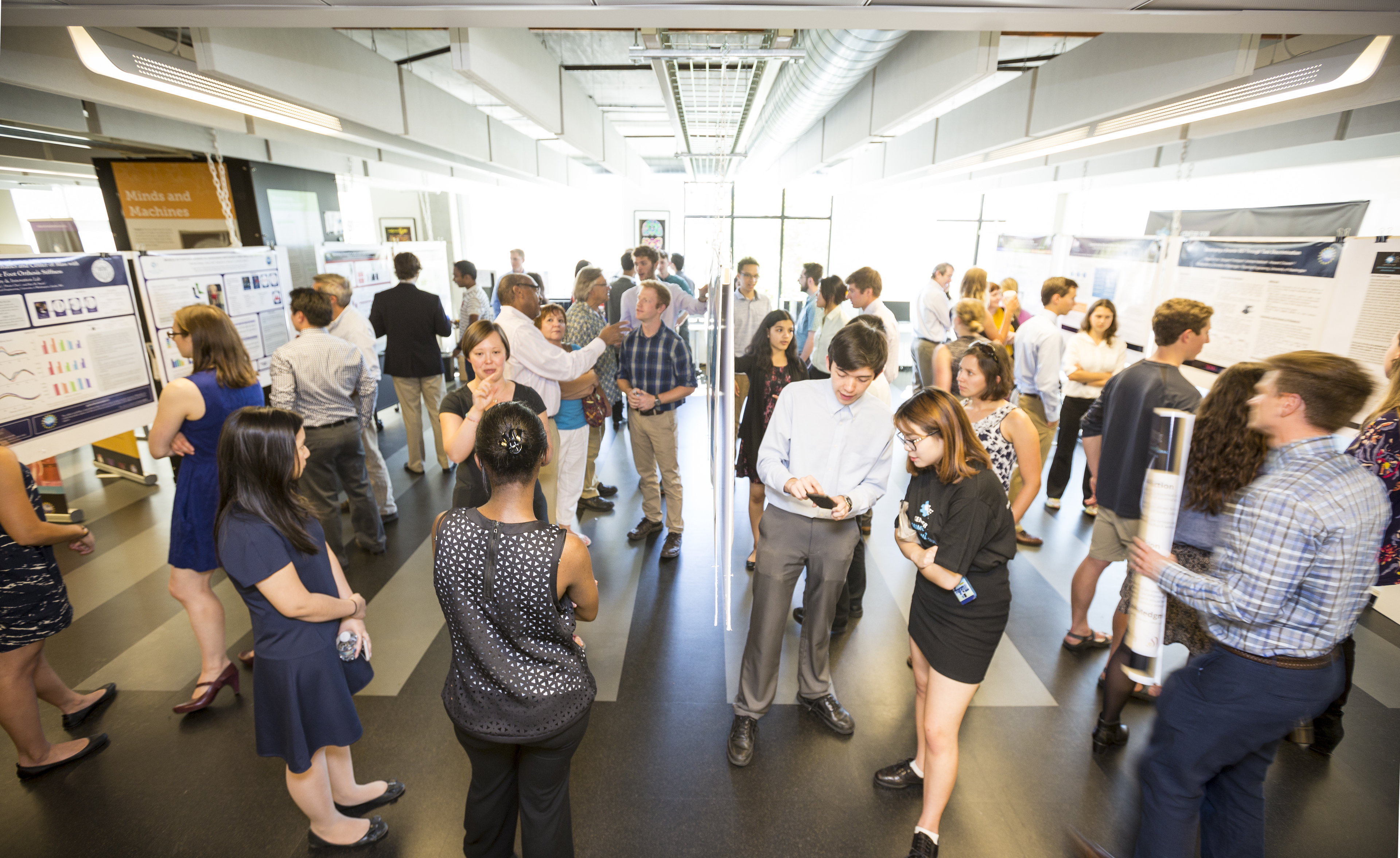 "A photo of a lively event, where people in business attire view posters lined up on rows of tables. Students, the creators of the posters, explain to the business people what the posters represent."